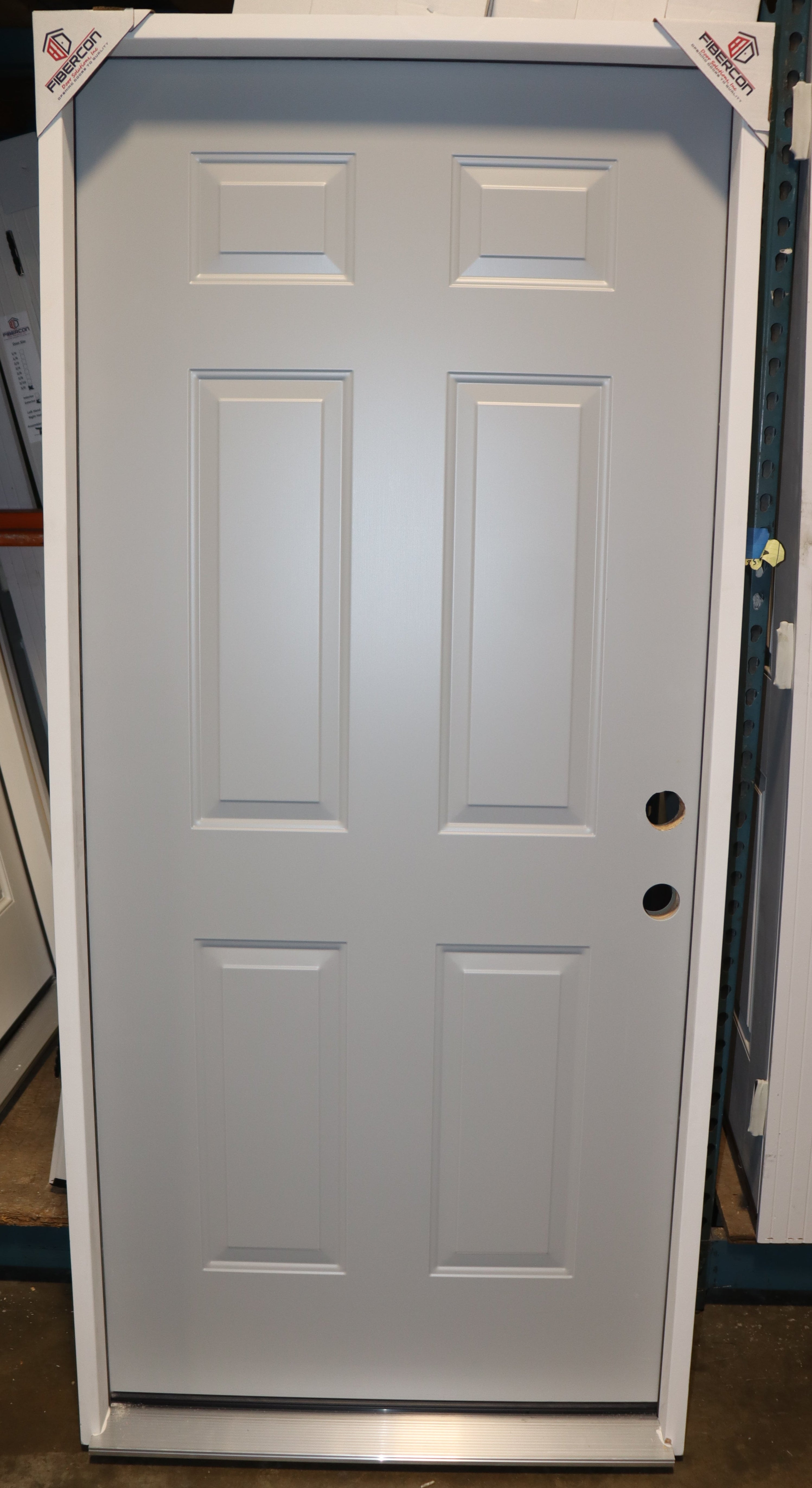 A white six-panel exterior door with black doorknobs, shown standing upright in a showroom or warehouse setting.