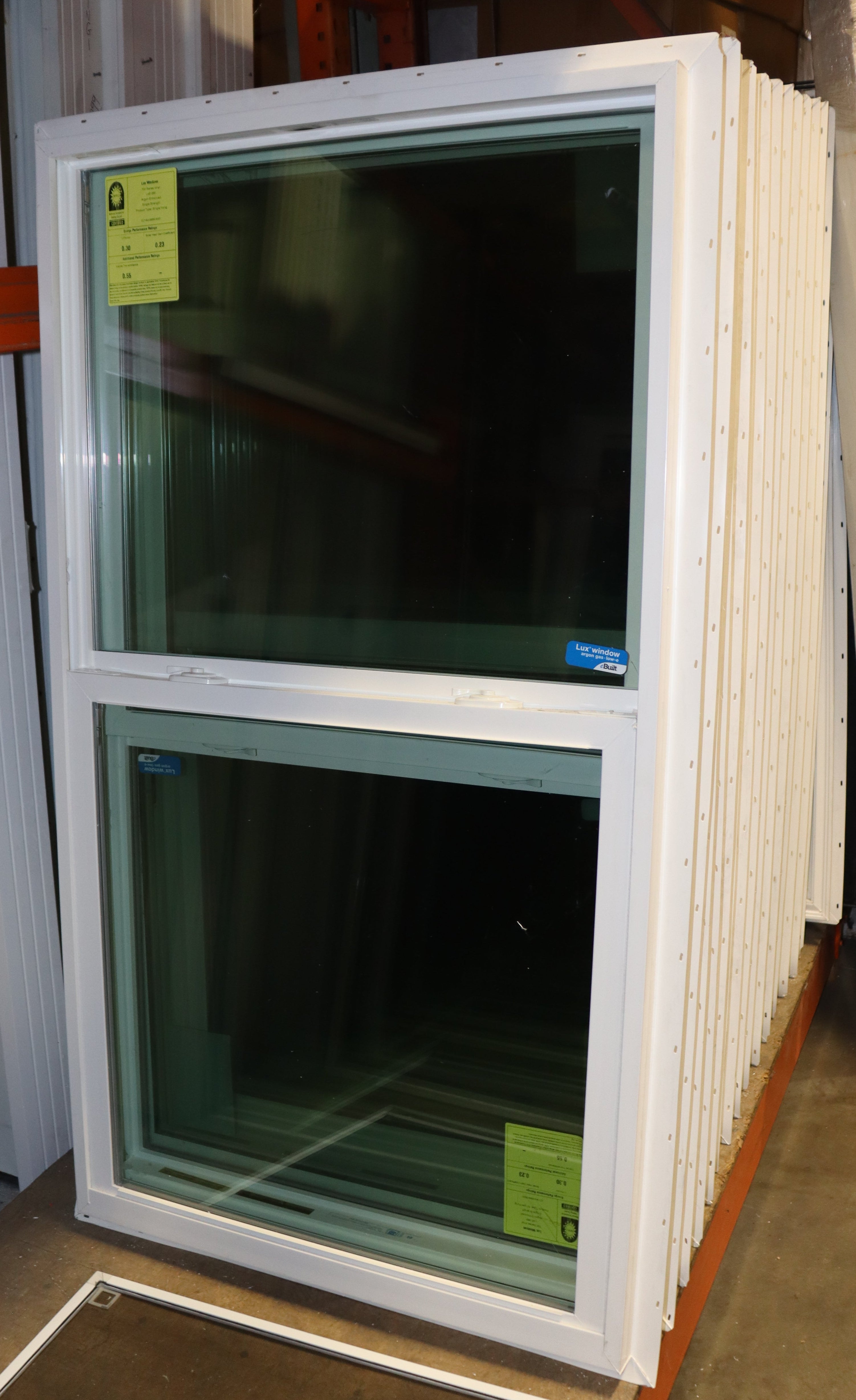 A white double-hung window unit with dark tinted glass stands upright in a warehouse or showroom, surrounded by stacks of similar window frames or panels.