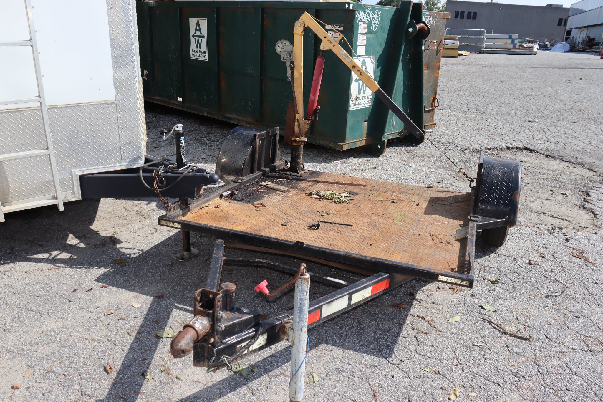 A used flatbed trailer with a rusty brown wooden deck, black metal frame, and hitch assembly, parked on a gravel lot next to a white trailer and a green dumpster.